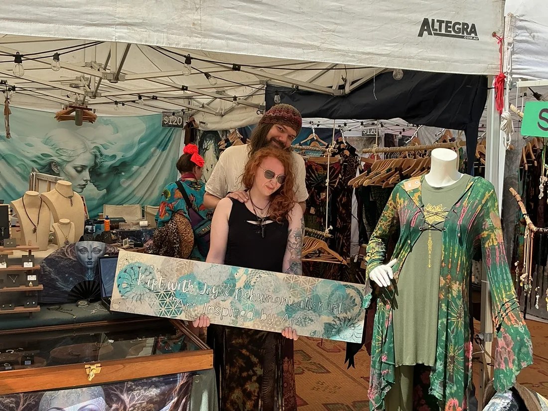 Charlie and Michael standing behind the Rapt with Joy festival stall sign with clothing, jewellery displays, and colourful tapestries.