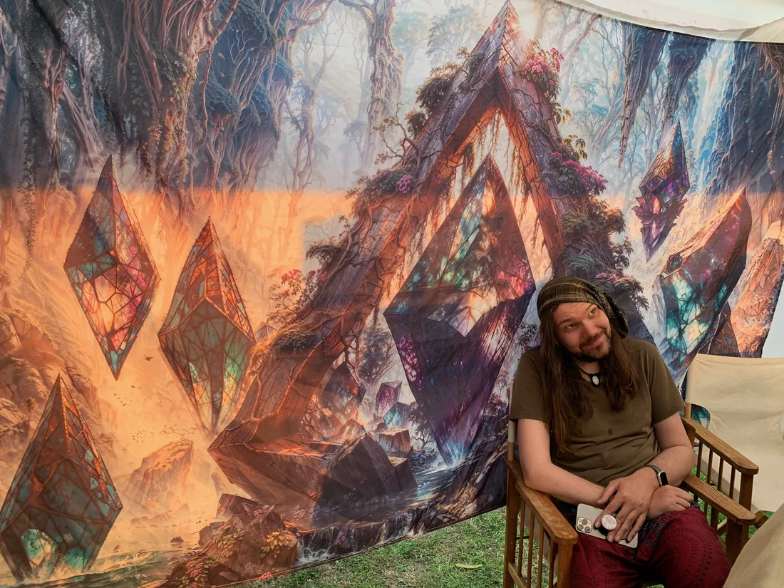 Michael sitting in front of a large colourful tapestry at the Rapt with Joy stall.